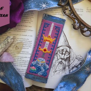 May include: A handmade bookmark with a cross-stitch design featuring a sword, crown, and a blue creature. The bookmark has a pink background with blue trim and the text "PDF PATTERN".