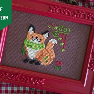 Op de afbeelding: Een kruissteekpatroon met een oranje vos die een groene sjaal draagt, op een donkerbruine stoffen achtergrond. Het ontwerp bevat de groene tekst "Joy" met hangende sterren en de rode tekst "25 AD", geplaatst in een rode lijst.