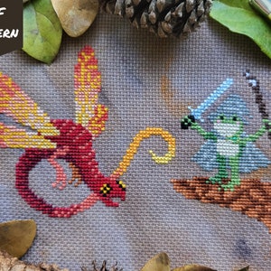 May include: Cross stitch pattern depicting a red and yellow dragon facing a green frog holding a sword and staff. The frog is wearing a blue robe and is perched on a brown branch. The background is a light grey fabric.