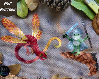 May include: Cross stitch pattern featuring a red and yellow dragon facing a green frog holding a sword and staff. The frog is wearing a blue robe and is sitting on a brown branch. The background is a light gray fabric.