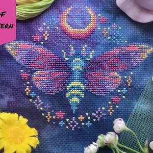 May include: A cross-stitch pattern featuring a celestial moth design. The moth has colorful wings with pink, purple, and blue hues, set against a dark blue background. A crescent moon and stars are above the moth. The words "PDF PATTERN" are in a pink box.