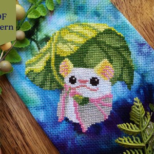 May include: Cross stitch pattern featuring a white mouse wearing a green and yellow leaf hat. The mouse is surrounded by a blue and purple watercolor background. The pattern is labeled "PDF Pattern".