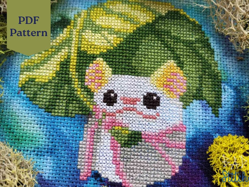 May include: Cross stitch pattern featuring a white cat with pink ears and a green leaf hat. The cat is wearing a pink scarf and is surrounded by a blue and green background.