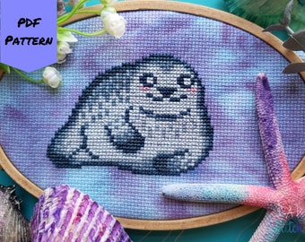 May include: Cross-stitch pattern of a cartoon seal with a blue and purple background. The pattern is framed in a wooden hoop.