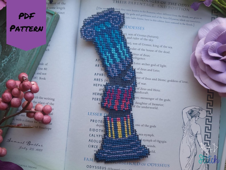 Op de afbeelding: Een handgemaakt kruissteek bladwijzer in een kolomontwerp, met tinten blauw, roze en geel. De bladwijzer ligt op een open boek met tekst en een paarse bloem op de achtergrond. De woorden "PDF PATTERN" staan op een paarse banner.
