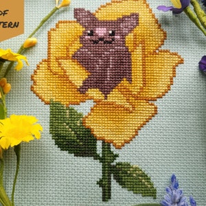 May include: Cross stitch pattern featuring a purple bat sitting inside a yellow flower with green leaves. The pattern is on a light blue background.