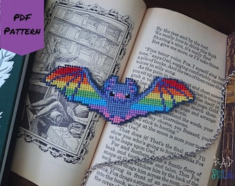 May include: A cross-stitch bat with rainbow wings and a blue body, resting on an open book. The bat is attached to a silver chain. The image also includes a purple tag that says "PDF PATTERN".