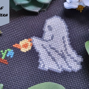 May include: Cross stitch pattern of a ghost holding a small plant with the word "yay" in colorful letters. The pattern is on black Aida cloth.