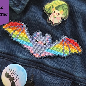 May include: A denim jacket adorned with a colorful, cross-stitched bat patch with rainbow wings. A clear pin featuring a cartoon animal with a leaf and a round button with a bat silhouette and the text "AUSTITCH" are also visible.