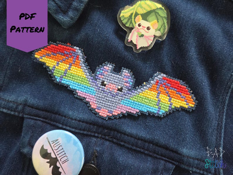 May include: A denim jacket adorned with a colorful, cross-stitched bat patch with rainbow wings. A clear pin featuring a cartoon animal with a leaf and a round button with a bat silhouette and the text "AUSTITCH" are also visible.