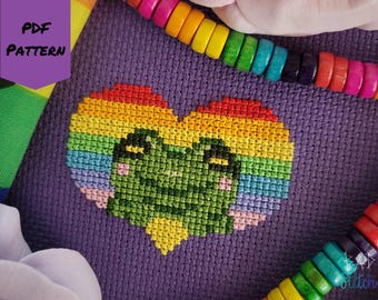 May include: Cross-stitch pattern featuring a rainbow heart with a frog face on a purple background. The heart is made of various colors, including red, orange, yellow, green, blue, and pink. The text "PDF PATTERN" is in a purple box in the upper left corner.