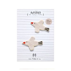 Könnte beinhalten: Zwei aufgestickte Vogelaufnäher mit cremefarbenen Körpern, roten Schnäbeln und schwarz-gelben Schwanzfedern. Auf einem gestreiften Hintergrund mit dem Text "AUFNÄHER" und "Anna Katharina Jansen Illustration".