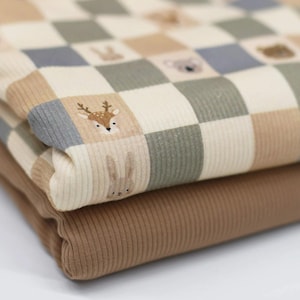 Könnte beinhalten: Ein gefaltetes Stück gerippter Stoff mit einem Karomuster in Creme, Beige, Salbeigrün und Schieferblau. Einige Quadrate enthalten kleine Illustrationen von Waldtieren, darunter ein Reh, ein Hase und ein Koala. Darunter liegt ein einfarbiger, brauner Rippstoff.