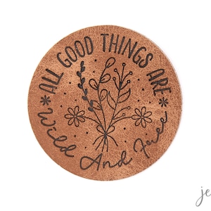 Könnte beinhalten: Ein brauner Lederuntersetzer mit dem Text "*All good things are wild and free*" in schwarzer Schrift. Der Text ist von einem floralen Design umgeben.