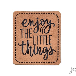 Könnte beinhalten: Ein rechteckiger, hellbrauner Aufnäher mit abgerundeten Ecken. Der Satz "enjoy the little things" ist in schwarzer Schreibschrift geschrieben. Der Aufnäher hat einen gepunkteten Rand und einen weißen Hintergrund.