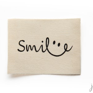 May include: A beige fabric label with the word "Smile" written in black cursive with a smiley face drawn next to it.