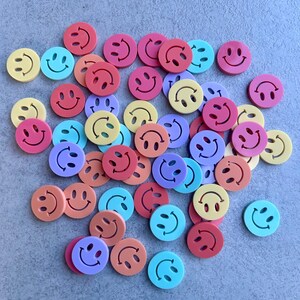 Könnte beinhalten: Eine Sammlung bunter Smiley-Knöpfe in verschiedenen Rosa-, Orange-, Gelb-, Blau- und Lilatönen. Jeder Knopf hat eine runde Form mit einem lächelnden Gesicht. Die Knöpfe sind auf einer grauen Oberfläche verstreut.