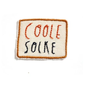 Könnte beinhalten: Ein weißer Stoff-Patch mit orangefarbenem Stickrand. Der Patch ist mit den Worten "COOL SOX" in orangefarbenem und schwarzem Garn bestickt.