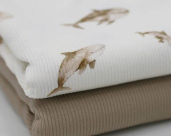 May include: Two folded pieces of fabric. The top layer is white with a repeating pattern of brown watercolor whales. The bottom layer is a solid tan color. Both fabrics have a ribbed texture.