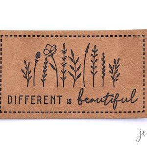 Könnte beinhalten: Ein braunes Leder-Patch mit einem genähten Rand. Das Patch zeigt eine schwarze Silhouette einer Reihe von Blumen und den Text "DIFFERENT is beautiful".