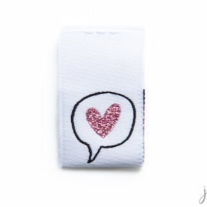 Könnte beinhalten: Weißes Stofflabel mit einem schwarzen Umriss einer Sprechblase, die ein pinkfarbenes Glitzerherz enthält.