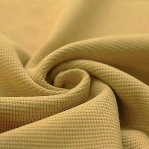 Könnte beinhalten: Nahaufnahme eines strukturierten, hellbraunen Stoffes mit einem waffelartigen Muster. Der Stoff ist gefaltet und drapiert, wodurch weiche Kurven und Schatten entstehen. Das Material scheint ein mittelschweres Strickgewebe zu sein, das für Kleidung oder Heimdekoration geeignet ist.