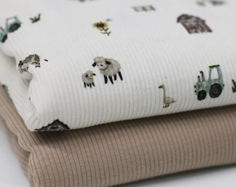 May include: Close-up of two folded ribbed fabrics. The top fabric is white with a repeating pattern of farm animals, tractors, and barns. The bottom fabric is a solid tan color. The fabrics are soft and appear to be cotton.
