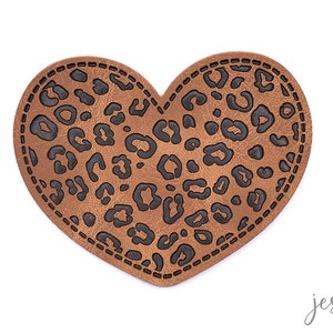Könnte beinhalten: Ein brauner herzförmiger Patch mit einem schwarzen Leopardenmuster. Der Patch hat einen gestickten Rand.
