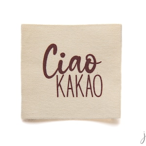 K&ouml;nnte beinhalten: Ein beigefarbenes Stofflabel mit dem Text "Ciao Kakao" in brauner Farbe.