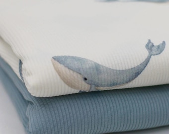 May include: Close-up of two folded fabrics. The top fabric is white with a ribbed texture and a watercolor whale print in shades of blue and beige. The bottom fabric is solid blue with a ribbed texture.
