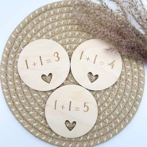 Könnte beinhalten: Drei runde Holzscheiben mit eingravierten mathematischen Gleichungen: 1+1=3, 1+1=4 und 1+1=5. Jede Scheibe hat einen herzförmigen Ausschnitt. Die Scheiben liegen auf einem geflochtenen beigen Platzset.
