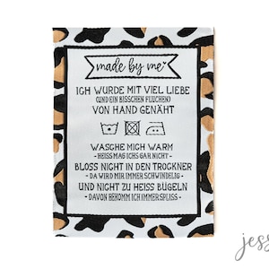 Könnte beinhalten: Ein weißes Stoffetikett mit schwarzem Text und einem Leopardenmuster-Rand. Das Etikett trägt die Aufschrift "made by me" und enthält Waschanweisungen. Der Text ist auf Deutsch, mit Sätzen wie "handgenäht" und "warm waschen".