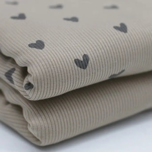 Könnte beinhalten: Beige gerippter Stoff mit einem sich wiederholenden Muster aus kleinen schwarzen Herzen. Der Stoff ist in der Hälfte gefaltet und zeigt die Textur des Materials.