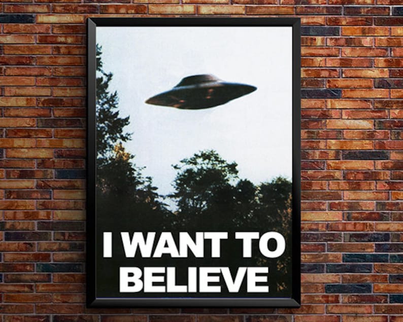 May include: A black framed poster with a white background and a black and grey UFO flying over a forest. The poster has the text "I WANT TO BELIEVE" in white letters.