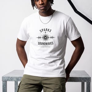 May include: A white short-sleeved t-shirt featuring a black graphic print on the chest. The design includes the text SPARKS & SOUNDWAVES, a central eye icon, and horizontal sound wave patterns. The shirt is paired with olive green cargo shorts.
