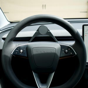 Puede incluir: Un volante de cuero negro con un logotipo de Tesla plateado en el centro. El volante tiene detalles plateados y botones en los lados izquierdo y derecho.
