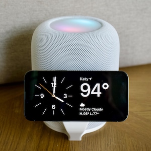 Può includere: Un altoparlante intelligente bianco con un design a rete, sormontato da una luce brillante. Uno smartphone nero mostra l'ora, una temperatura di 34 gradi Celsius e le condizioni meteorologiche. Il telefono è su un supporto bianco.