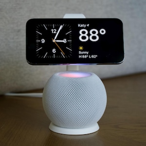 May include: A white smart speaker with a white charging stand and a smartphone on top. The smartphone screen displays a clock, the temperature, and the word "Sunny".