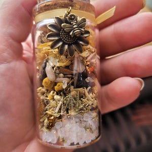 May include: A small glass jar with a cork lid and a metal charm of a sunflower with a bee. The jar is filled with dried herbs, flowers, and crystals.