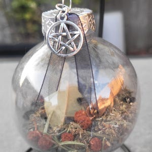 May include: A clear glass bauble filled with dried herbs, berries, and a small piece of paper. A silver pentagram charm hangs from the top, along with a black ribbon. The bauble is designed for decorative purposes.