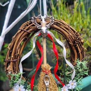 May include: A small wreath made of brown twigs with white and red ribbons, a small broom with a silver pentacle charm, and a silver stag head charm.