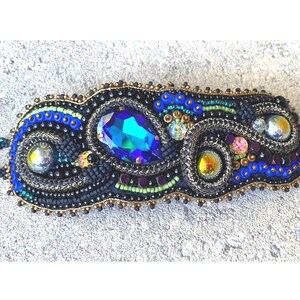 May include: A beaded bracelet with a black base and gold accents. The bracelet features a large iridescent blue gemstone and smaller iridescent beads. The bracelet is designed in a swirling pattern.