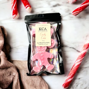 May include: A package of pink soy wax melts in the shape of a gingerbread person. The package is labeled "KCA Candy Cane Soy Blend Wax Melts 103 g Orangville, ON, Canada".