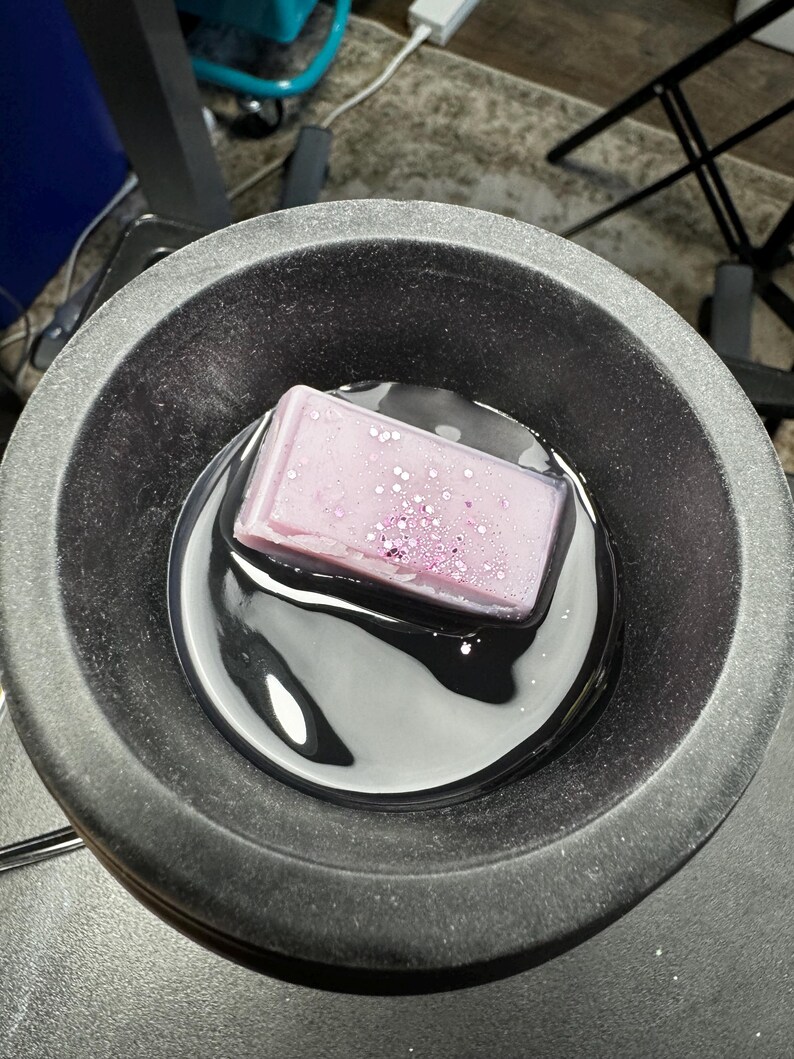 May include: A purple wax melt with glitter is melting in a black wax warmer.