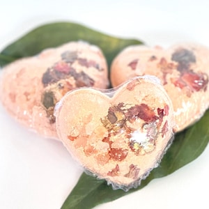 May include: Three heart-shaped bath bombs, wrapped in clear plastic, are displayed on a green leaf against a white backdrop. The peach-coloured bath bombs are decorated with dried flower petals.