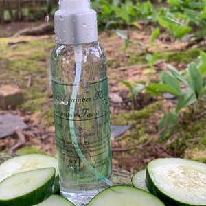 May include: A clear glass bottle with a white spray top and a green label that reads "Cue-cumber Refresh Cucumber Facial Toner". The bottle is sitting on a wooden surface with sliced cucumbers.