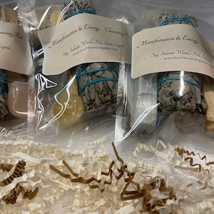May include: Three clear plastic bags containing smudging kits. Each kit includes a bundle of white sage, a palo santo stick, and a crystal. The bags are labeled with the text "Energy Cleansing Kit", "Manifestation & Energy Cleansing Kit", and "Manifestation & Energy Cleansing Kit".