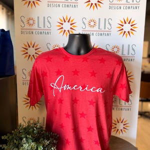 May include: A red short-sleeved t-shirt featuring a repeating pattern of darker red stars across the fabric. The word America is printed across the center of the chest in a white cursive script font. The shirt is displayed on a black mannequin torso.