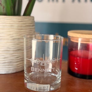 May include: A clear glass tumbler with the name "BENNETT" etched on the front, along with an airplane design. The glass is cylindrical and sits on a wooden surface. A red candle in a glass jar is visible in the background.