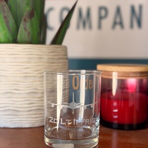 May include: A clear glass tumbler with an etched airplane design and the text "2D LT IF RIG". A red candle in a black jar and a potted plant are in the background.